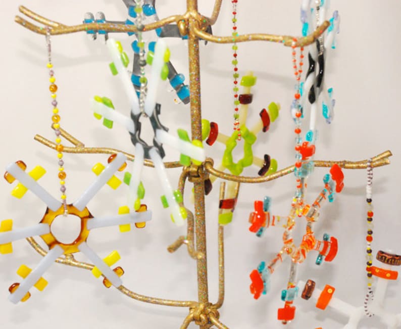 May include: A gold metal stand with multiple branches holds a collection of colorful glass snowflake ornaments. The snowflakes are made of various colors, including red, orange, yellow, green, blue, and white. Each snowflake is unique in its design and shape.