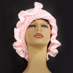Close-up of a pink crochet ruffle hat with layered petal design, showcasing the intricate texture and elegant Mobius twist, modeled on a mannequin head against a black backdrop