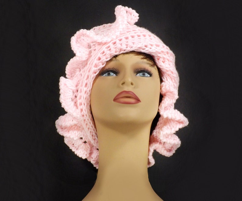 Close-up of a pink crochet ruffle hat with layered petal design, showcasing the intricate texture and elegant Mobius twist, modeled on a mannequin head against a black backdrop