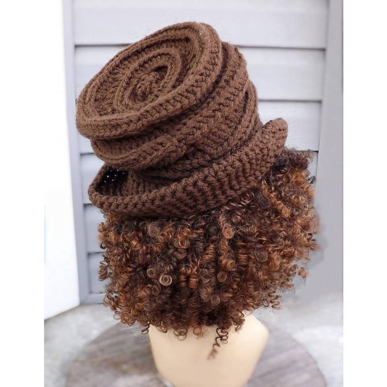 A rear view of a brown crocheted hat with a swirled, ribbed texture on a mannequin head. The hat sits tall and has tight curls of brown hair hanging down the back and sides of the head.