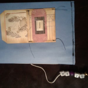 May include: A blue notebook with a sewn edge and a pocket on the front. The pocket contains a piece of paper with a feather design and a pink card with the word "Notes" written on it. A white string with beads that spell "XYZG" is attached to the notebook.
