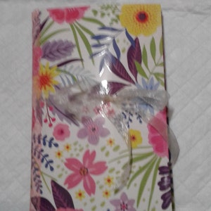 May include: A white journal with a floral design featuring pink, yellow, and purple flowers. The journal is tied with a white ribbon.