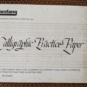May include: A white practice paper pad for calligraphy. The pad has a slanted grid and a practice rule. The text on the front of the pad reads "Calligraphic Practice Paper" and "Bienfang Calligraphic Practice Paper No. 206". The pad contains 50 sheets and measures 12 x 9 inches.