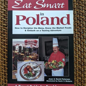 May include: A travel guide titled "Eat Smart in Poland" with a black cover and red and white text. The cover features images of Polish food and a chef. The book promises to help readers decipher menus and explore Polish cuisine.