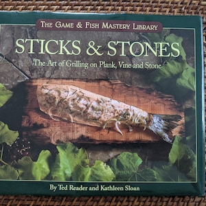 May include: A cookbook titled "STICKS & STONES" from The Game & Fish Mastery Library. The cover features a grilled fish on a wooden plank, surrounded by green leaves. The book's subtitle is "The Art of Grilling on Plank, Vine and Stone."