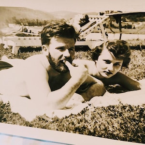 May include: A vintage sepia-toned photograph showing two individuals lying on a blanket outdoors. The person on the left has a beard and is touching their mouth with a finger. The person on the right has dark wavy hair and is looking forward.