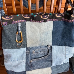 以下が含まれることがあります： パッチワークデニムのトートバッグ。黒い模様のトップエッジとストラップが付いています。バッグは、さまざまな色合いの青いデニムの正方形、金のカラビナクリップ、デニムのポケットが特徴です。バッグは木製の椅子の上に置かれています。