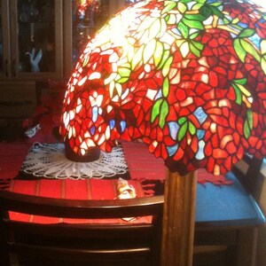 May include: A stained glass floor lamp with a red, green, and blue floral design. The lamp is on a wooden base and has a white doily on the table below it.