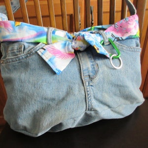 May include: A blue denim tote bag with a colorful fabric strap. The strap is tied in a bow at the front of the bag. A green carabiner clip is attached to the strap.