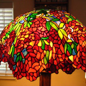 May include: A stained glass lampshade with a floral design. The shade is made up of many small pieces of glass in shades of red, orange, yellow, green, and blue. The glass is arranged in a pattern of flowers and leaves.