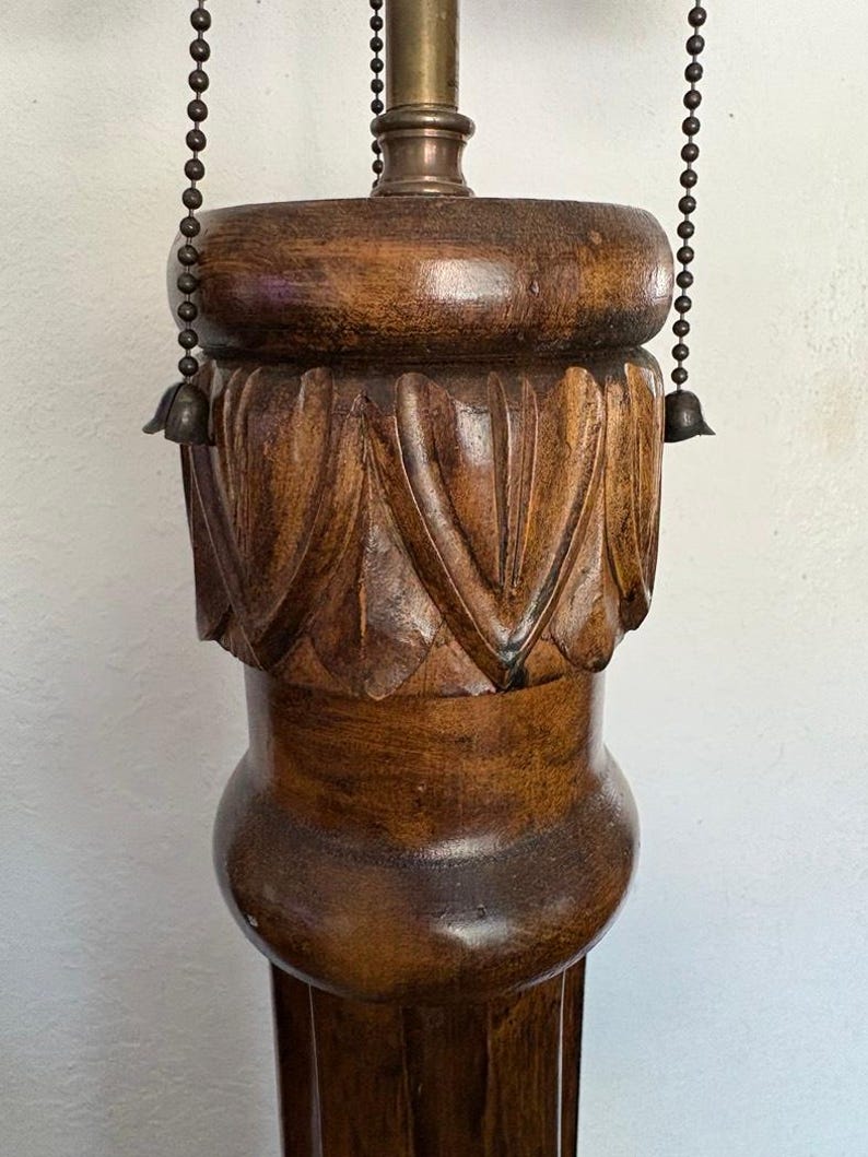 May include: A tall, wooden lamp base with a carved design. The base is dark brown and has a fluted column. The lamp has a chain for adjusting the height of the shade.