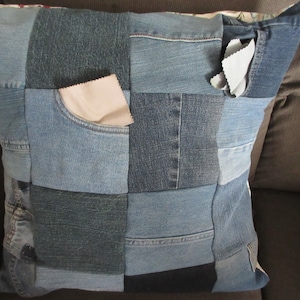 May include: A square pillow cover made from patchwork denim fabric. The pillow cover is made from different shades of blue denim and has a few pockets.