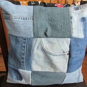 May include: A patchwork pillow made from recycled denim jeans. The pillow features various shades of blue denim, including light and dark washes, with a pocket detail. The top edge has a black fabric with a floral pattern.