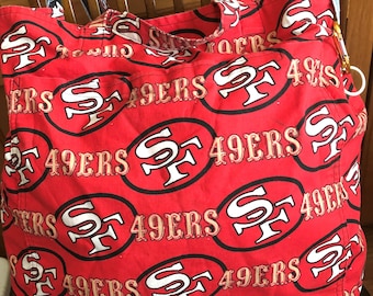 Puede incluir: Bolsa de tela roja con un patrón de logo de los San Francisco 49ers en blanco y dorado. La bolsa tiene asas de mezclilla.