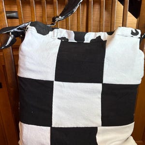 Könnte beinhalten: Eine schwarz-weiße Tragetasche mit Schachbrettmuster auf der Vorderseite. Die Tasche hat einen langen schwarzen Riemen und einen oberen Bereich mit Kuhmuster. Die Tasche liegt auf einem Holzstuhl.