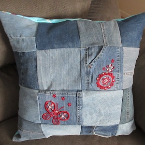 May include: A blue denim pillow with red embroidered butterflies and flowers. The pillow is made from recycled denim and has a patchwork design.
