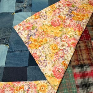 May include: A patchwork quilt with a denim base and a floral patterned overlay. The denim sections are various shades of blue. The floral fabric features pink, yellow, and orange flowers. The quilt is a mix of textures and colors.