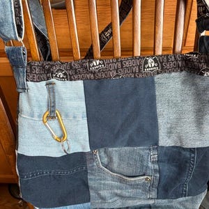Könnte beinhalten: Eine Patchwork-Denim-Tragetasche mit Raiders-Logo-Gurt und goldenem Karabinerhaken. Die Tasche besteht aus verschiedenen Blautönen von Denim, einschließlich einer Taschendetail. Der Gurt zeigt den Text "RAIDERS" wiederholt.