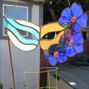 May include: A stained glass mask with a yellow and white design and blue flowers. The mask is hanging from a chain.