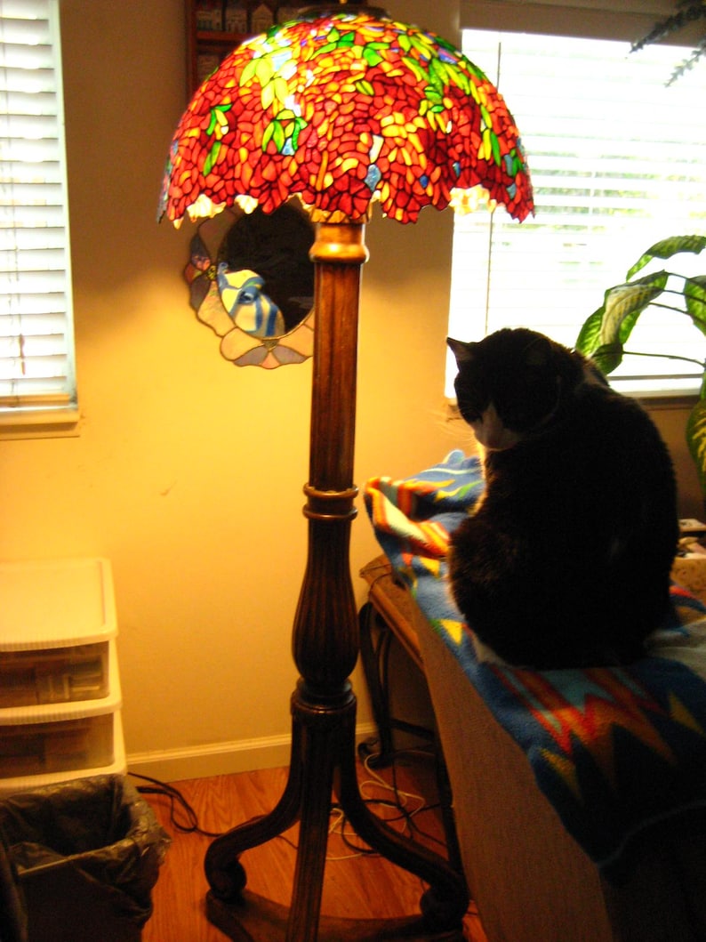 May include: A stained glass lamp with a floral design in shades of red, orange, and yellow. The lamp is on a wooden base and has a black cat sitting on a blanket nearby.