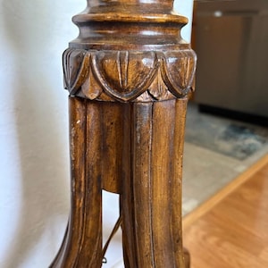 May include: Close-up of the base of a wooden floor lamp. The base is intricately carved with a fluted design and a decorative band of leaves. The lamp is made of dark brown wood and has a traditional style.
