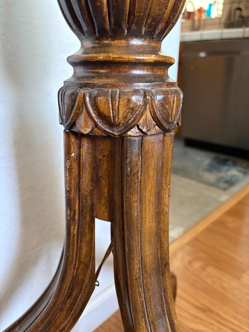 May include: Close-up of the base of a wooden floor lamp. The base is intricately carved with a fluted design and a decorative band of leaves. The lamp is made of dark brown wood and has a traditional style.