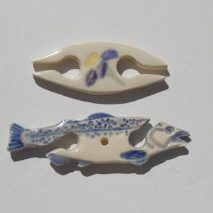 May include: Two ceramic toggles. The top toggle is an off-white almond shape with a painted blue and yellow flower. The bottom toggle is shaped like a fish with blue and white speckled details, a blue tail, and a central hole for threading.