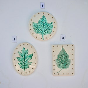 May include: Three ceramic leaf-shaped ornaments. The ornaments are in a cream color with a green leaf design in the center. Each ornament has a different shape: round, oval, and rectangular. Small brown dots border each ornament.