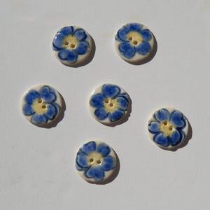 May include: A set of five round ceramic buttons, each featuring a hand-painted blue flower design with a yellow center. Every button has two small holes in the middle for attachment, with a smooth, off-white base finish throughout.