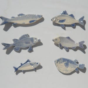 May include: A collection of six ceramic fish ornaments, each featuring a unique shape and blue and white hand-painted details. The set includes various species with distinct fin structures, scale patterns, and facial features, all finished with a glossy glaze.