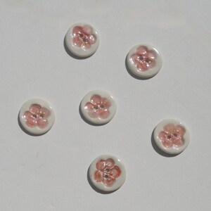 May include: Six circular white buttons, each featuring a raised, hand-painted pink flower design with a glossy finish. Every button has two small holes in the center for attachment, arranged in a scattered pattern across the frame.