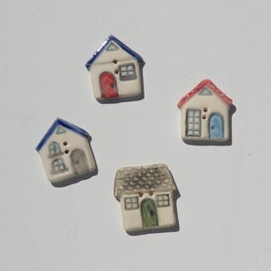 May include: Four ceramic buttons shaped like small houses. Each button features a unique painted design with details including blue, red, or green doors, windows, and roofs. The buttons have two small holes in the center for attachment.