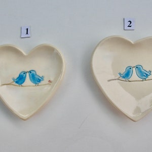 May include: Two blue birds are kissing on a branch, painted on a white heart-shaped ceramic dish. The dish is approximately 4 inches in diameter.