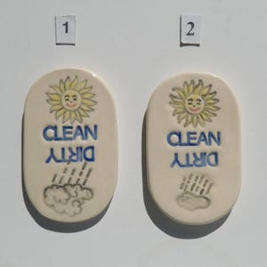 May include: Two ceramic dishwasher magnets with a cream background. Each magnet has a smiling sun, the words "CLEAN" and "DIRTY" in blue, and a cloud with rain. The magnets are oval-shaped.