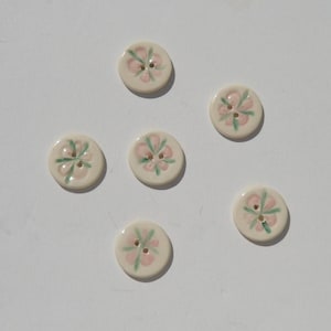 May include: Five round, off-white ceramic buttons, each featuring a hand-painted floral design with soft pink petals and green leaves. Every button has two small holes in the center for attachment, showcasing a smooth, glossy finish throughout.