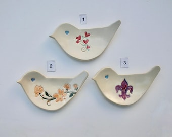 May include: Three off-white ceramic bird-shaped dishes. One dish has red heart accents, another has peach-colored floral designs, and the third features a purple fleur-de-lis. Each dish has a blue heart detail.