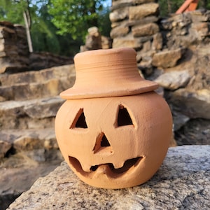 May include: A terracotta pumpkin-shaped candle holder with a removable lid. The holder has a carved jack-o'-lantern face with triangular eyes, a wide nose, and a smiling mouth.