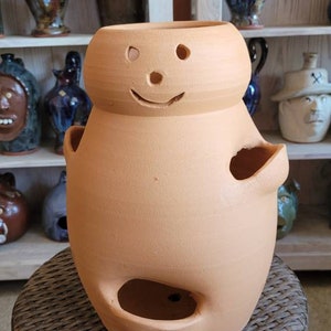 May include: A terracotta-colored ceramic planter with a smiling face design. The planter has two arm-like handles and a large opening at the bottom. It is sitting on a woven wicker stool, with other ceramic items on shelves in the background.