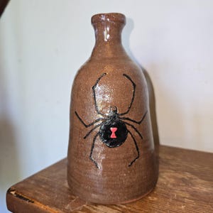 May include: A brown ceramic bottle with a glossy finish featuring a hand-painted black widow spider on the front. The spider is depicted in black with a distinct red hourglass marking on its abdomen. The bottle has a narrow neck and a wide base.