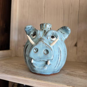 May include: A light blue ceramic piggy bank shaped like a wild boar with two prominent white tusks protruding from the snout. The glaze features dark brown accents around the ears, eyes, and snout, with a small circular opening at the top center.