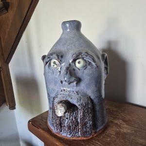 Könnte beinhalten: Ein Keramik-Gesichtskrug mit glänzender, kornblumenblauer Glasur, der ein skulptiertes menschliches Gesicht mit weit aufgerissenen Augen, einer markanten Nase und einem texturierten, dunkelbraunen Bart zeigt. Ein zylindrisches Objekt ragt aus dem Mund.