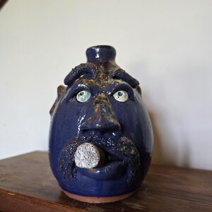 May include: A dark blue glazed ceramic face jug featuring a sculpted nose, thick textured eyebrows, and a prominent mustache. The figure has wide, light-colored eyes and a small white cylindrical object protruding from the mouth area.