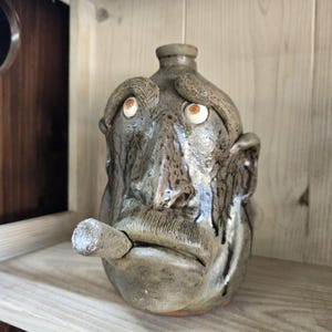 May include: A ceramic jug shaped like a human head with a mottled brown and grey glaze. The face features a prominent nose, thick eyebrows, large ears, and a mustache. A cylindrical object resembling a cigar protrudes from the mouth area.