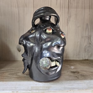 May include: A dark metallic gray ceramic jug featuring a sculpted face with multiple protruding eyes, a textured circular object held in the mouth, and two prominent white teeth. The vessel has a smooth, glossy finish and a curved handle at the top.