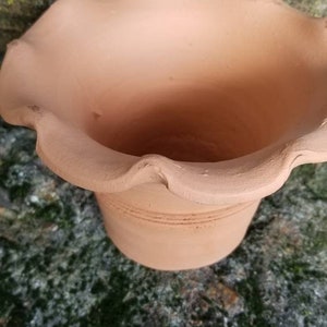 May include: A terracotta-colored ceramic pot with a ruffled rim. The pot has a cylindrical base with horizontal etched lines. The pot is sitting on a mossy, textured surface.