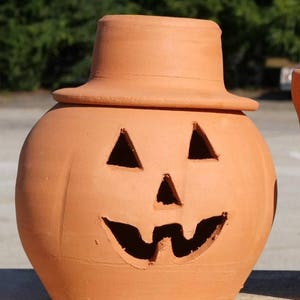 May include: A terracotta pumpkin-shaped container with a hat-shaped lid. The pumpkin has triangular eye cutouts and a carved, smiling mouth. The container is a warm orange color, perfect for Halloween decorations.
