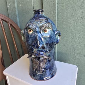 May include: A ceramic jug shaped like a human head with a glossy dark blue glaze. The face features textured eyebrows, a mustache, and a goatee. The eyes are round with brown irises. The jug has a narrow neck opening at the top of the head.