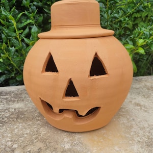 May include: A terracotta jack-o'-lantern sculpture featuring a round body with cut-out triangular eyes and nose, a jagged mouth, and a matching terracotta hat lid resting on top. The surface is matte, unglazed, and light orange in color.