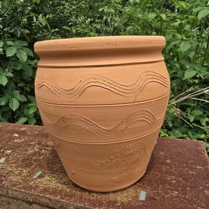 May include: A large, unglazed terracotta pot with a wide rim and a bulbous body. The pot features horizontal bands with carved, wavy lines. The natural clay color complements the outdoor setting, suggesting a garden or patio item.