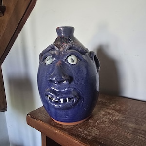 May include: A dark blue glazed ceramic face jug featuring a sculpted human-like visage with prominent eyebrows, wide eyes, a nose, and a mouth displaying small white teeth. The vessel has a narrow neck opening at the top and a rounded base.